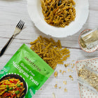 Amritatva gluten-free multigrain pasta packaging with pasta on a plate and around it on a wooden surface.